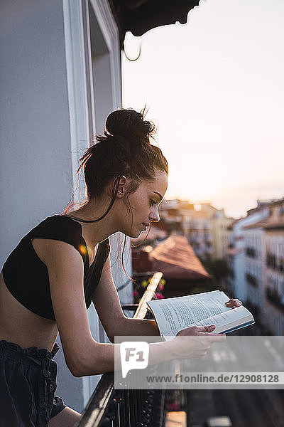 Beautiful young woman on balcony above the city at sunset reading a book