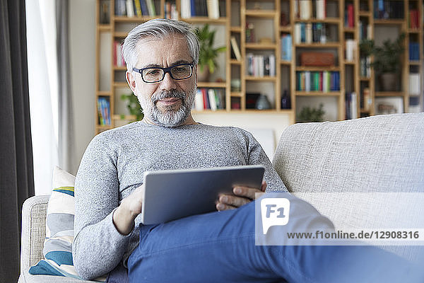 Portrait of mature man sitting on couch at his living room using tablet