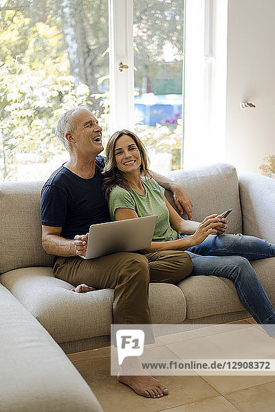 Happy mature couple sitting on couch at home with cell phone and laptop