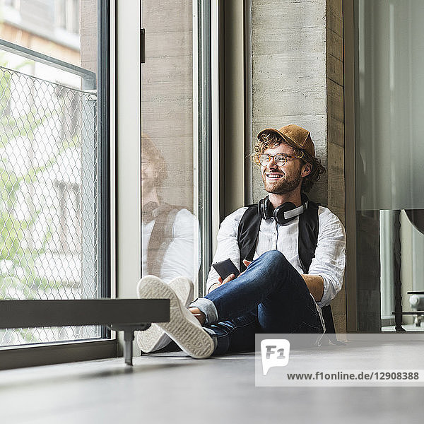 Smiling casual young man with headphones and cell phone sitting at the window