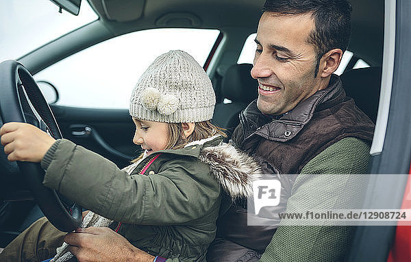 Happy little girl with wool cap driving car sitting on father's lap