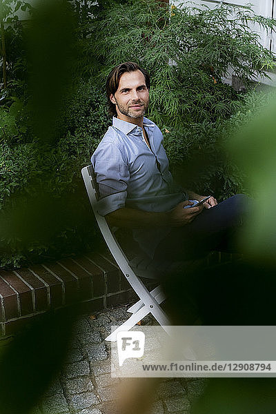 Portrait of smiling man sitting on chair in garden using cell phone