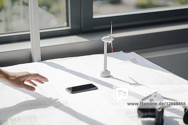 Hand of man reaching for smartphon lying on desk with blueprints and wind wheel