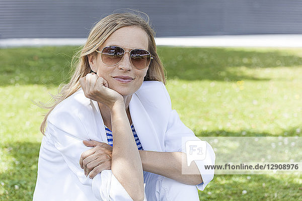 Smiling blond businesswoman sitting on meadow  hand on chin