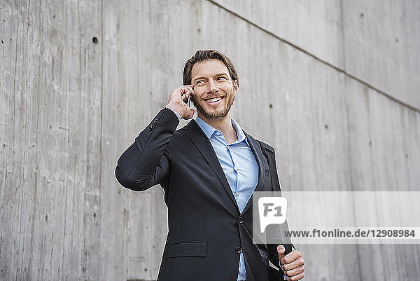 Smiling businessman at concrete wall talking on smartphone