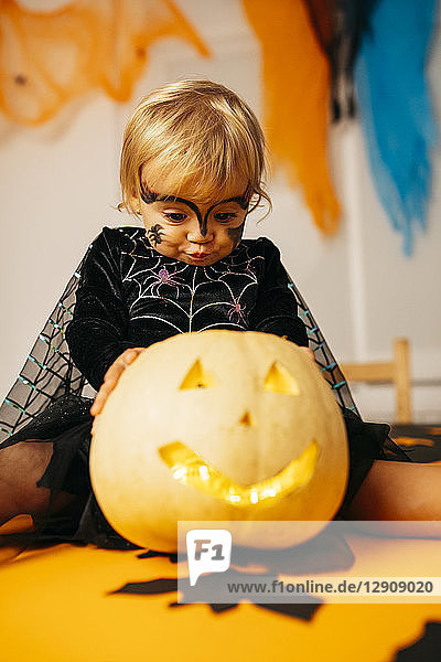 Portrait of little girl with painted face and fancy dress sitting on table with Jack O'Lantern pouting mouth