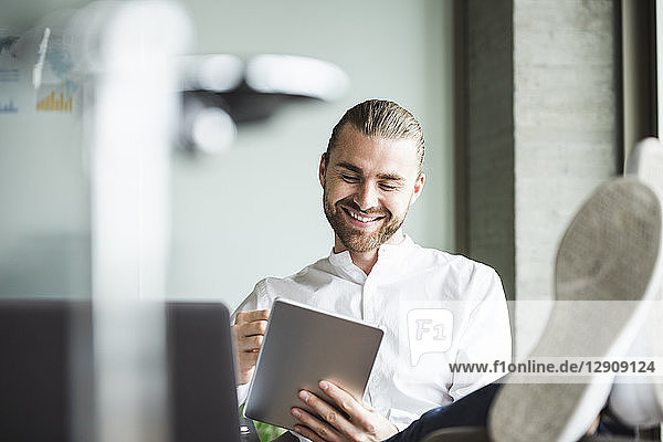 Smiling businessman sitting in office with feet up using tablet