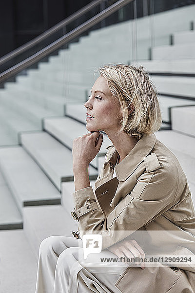 Young blond businesswoman wearing beige trenchcoat sitting on stairs