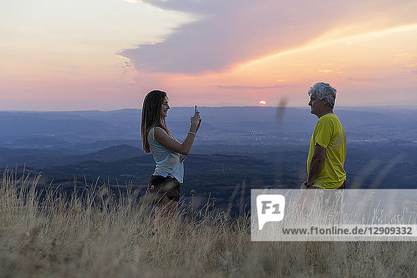 Spain  Catalonia  Montcau  senior father and adult daughter taking a cell phone picture on top of hill during sunset