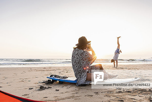 Young woman sitting on surfboard  taking pictures of young man  practicing handstands on the beach