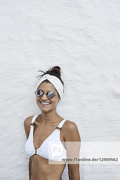 Portrait of laughing young woman wearing white bikini top leaning against white wall