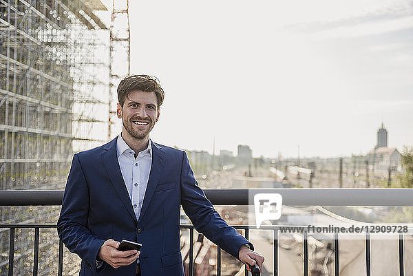 Portrait of smiling businessman standing on bridge in the city holding cell phone