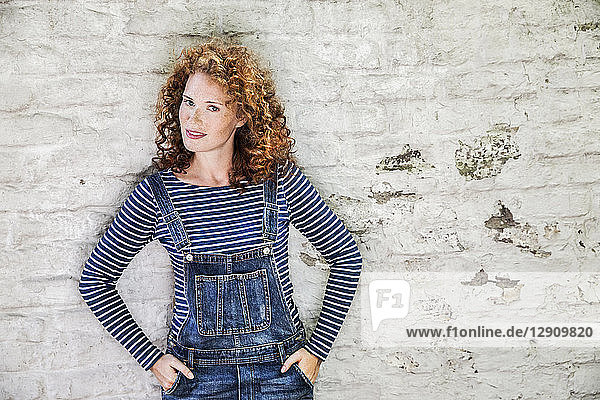 Portrait of smiling young woman wearing denim dungarees leaning against white brick wall
