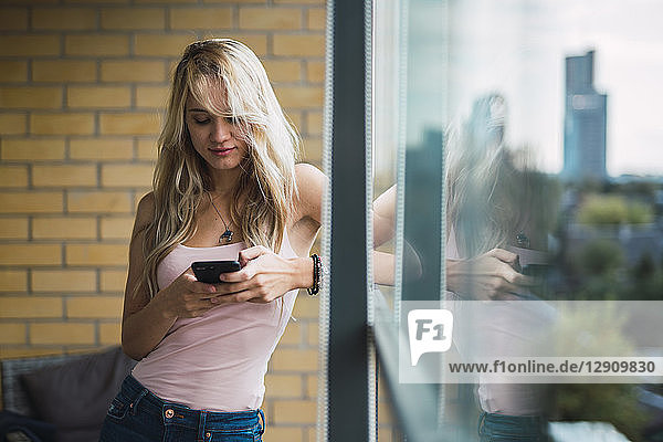 Blond young woman checking cell phone at the window
