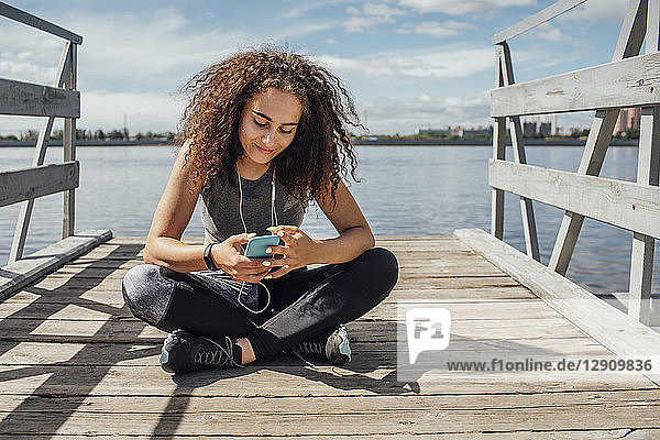 Young athletic woman sitting at the riverside using smartphone
