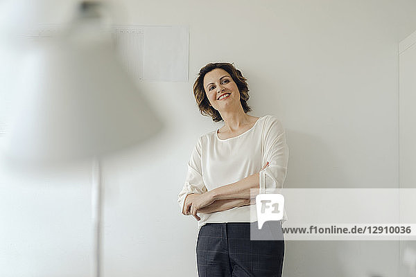 Businesswoman leaning against wall in office  with arms crossed  smiling