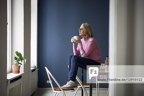 Mature woman at home with cup of coffee looking out of window