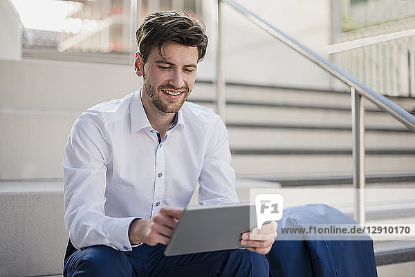 Smiling businessman sitting on stairs in the city using tablet