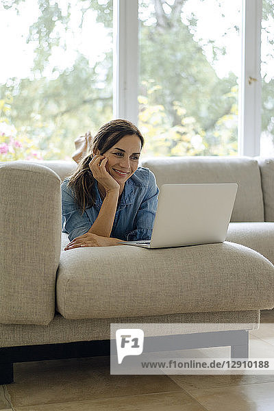 Smiling mature woman lying on couch at home using laptop