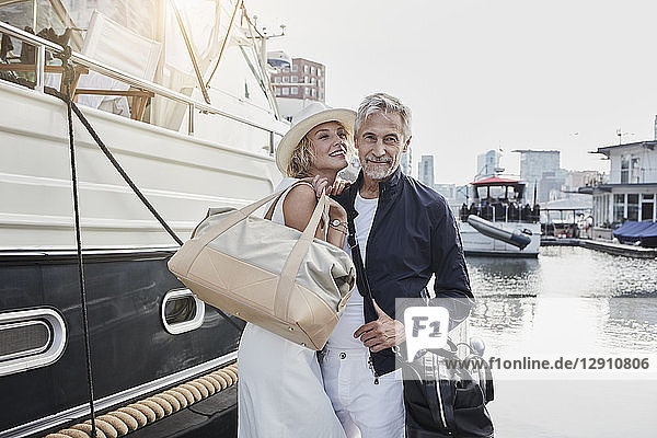 Older man and young woman standing with travelling bags on jetty next to yacht