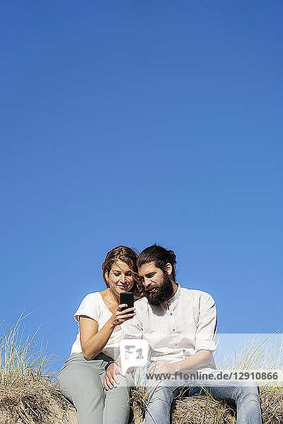 Young couple sitting on a dune in summer  using smartphone