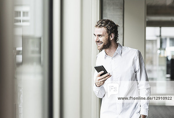 Smiling young businessman holding cell phone looking out of window in office