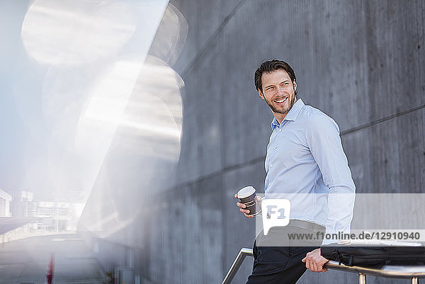 Smiling businessman with laptop bag and takeaway coffee leaning against a railing