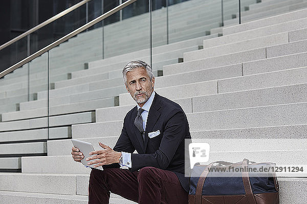Portrait of fashionable businessman with travelling bag and tablet sitting on stairs