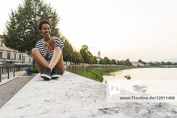 Smiling young woman sitting on wall at the riverside using cell phone