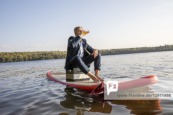 Businessman sitting on surfboard on a lake having a drink