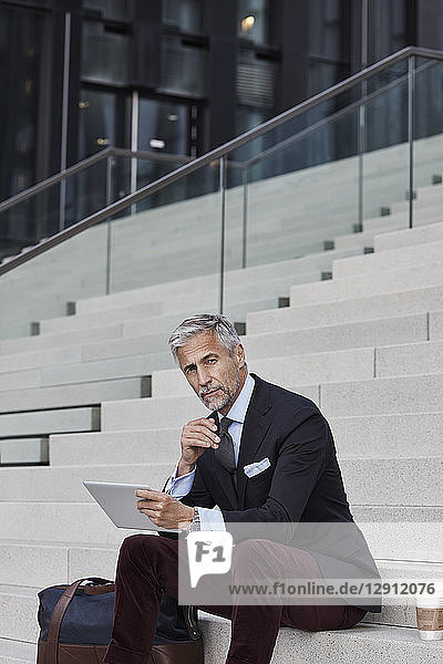 Portrait of fashionable businessman with travelling bag  coffee to go and tablet sitting on stairs
