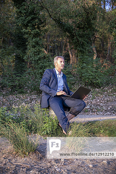 Mature businessman with laptop sitting barefoot in nature looking at distance