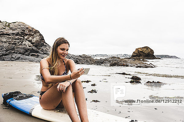 Young woman with surfboard sitting on the beach  using smartphone
