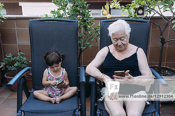 Grandmother and baby girl sitting side by side on the terrace using mobile phones
