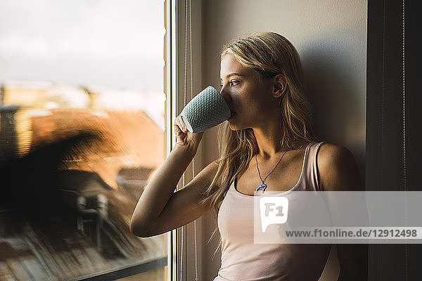 Blond young woman drinking coffee from mug looking out of window