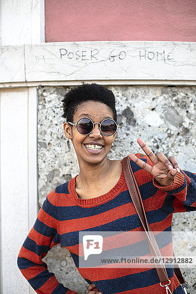 Portrait of smiling young woman wearing sunglasses and striped pullover showing victory sign