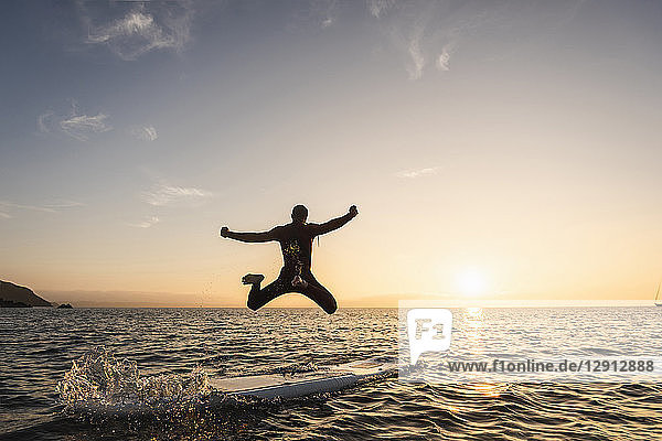 Young man jumping from paddleboard into water at sunset