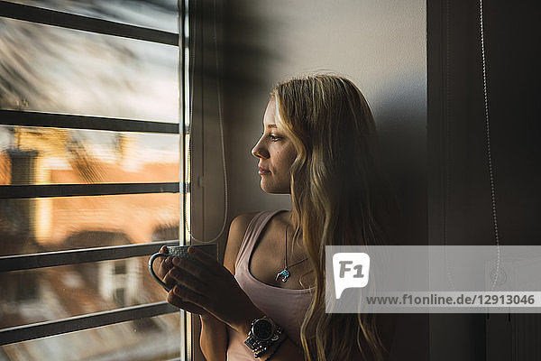 Blond young woman holding coffee mug looking out of window