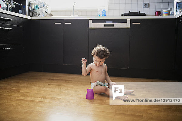 Baby boy sitting on floor in the kitchen playing with plastic cup