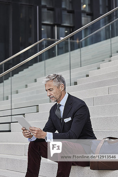 Fashionable businessman with travelling bag sitting on stairs using tablet