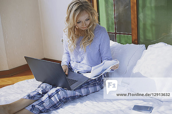 Woman sitting on bed with laptop  reading papers