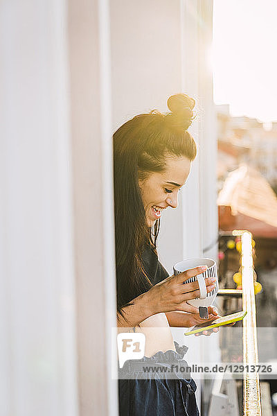 Happy young woman on balcony above the city at sunset holding cell phone and cup of tea