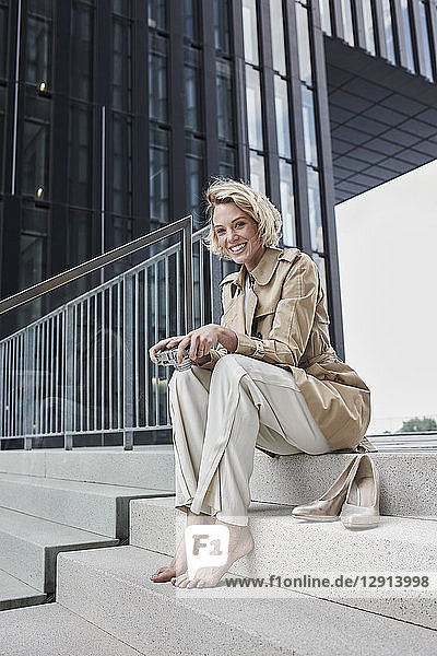 Portrait of laughing young blond woman with digital camera sitting barefoot on stairs