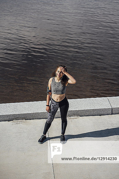 Exhausted young athletic woman wearing earbuds standing at the riverside