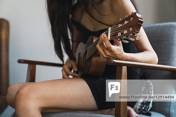 Young woman sitting on armchair at home playing guitar
