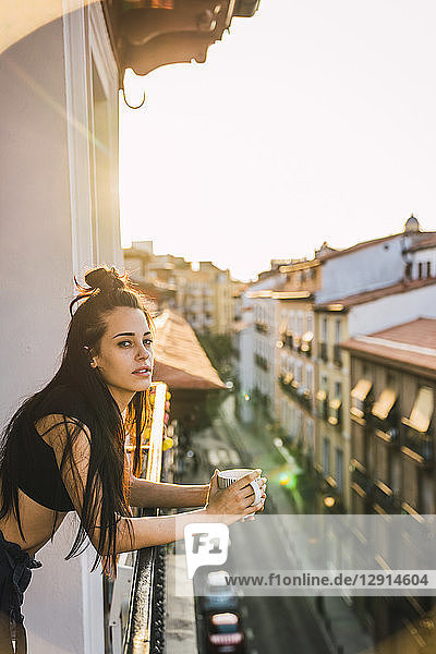 Beautiful young woman on balcony above the city at sunset with cup of coffee