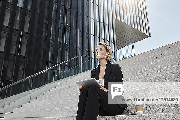 Blond businesswoman with tablet and coffee to go sitting on stairs in front of modern office building