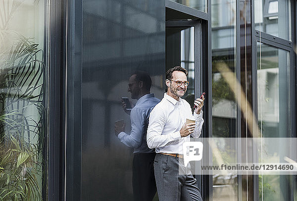 Businessman with coffee to go using cell phone in front of office building