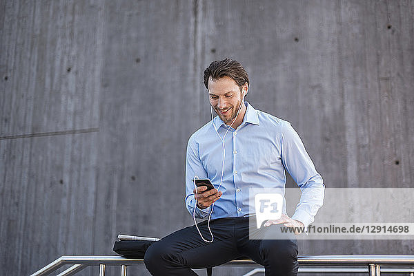 Smiling businessman with earbuds sitting on a railing looking at smartphone