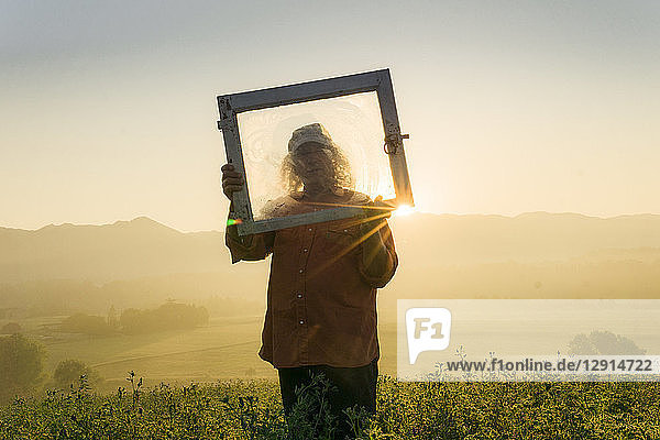 Italy  Tuscany  Borgo San Lorenzo  senior man holding window frame in field at sunrise above rural landscape